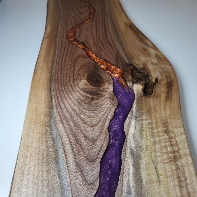 May include: A wooden serving board with a natural wood grain finish. A purple and gold resin river runs through the center of the board.