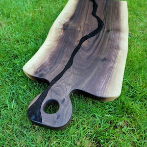 May include: A large, dark wood serving board with a black epoxy river running through the center. The board has a unique, hand-shaped handle with a hole in the center.