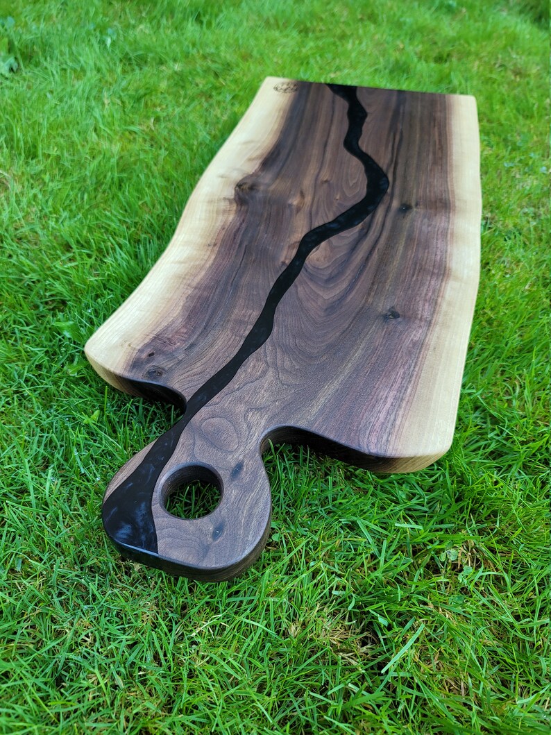 May include: A large, dark wood serving board with a black epoxy river running through the center. The board has a unique, hand-shaped handle with a hole in the center.