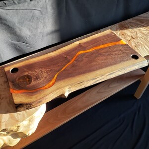 May include: A large wooden serving board with a natural edge and a dark brown finish. A bright orange resin river flows through the center of the board.