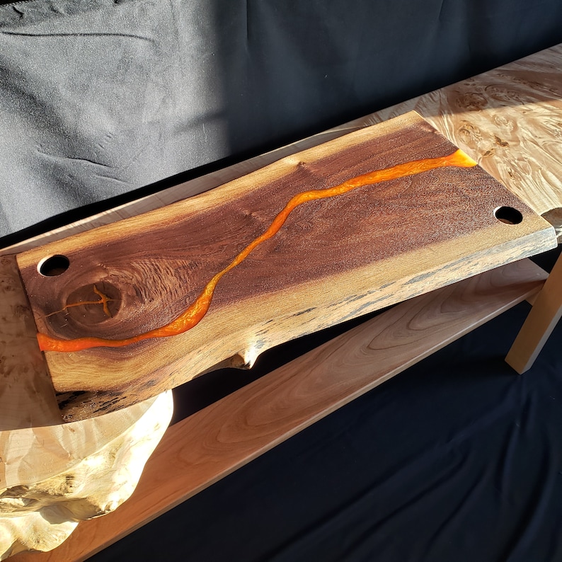 May include: A large wooden serving board with a natural edge and a dark brown finish. A bright orange resin river flows through the center of the board.