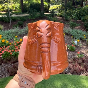 May include: A brown ceramic mug with an elephant design. The mug has an elephant trunk handle and embossed facial features. The mug is held in front of a blurred outdoor background with trees and flowers.
