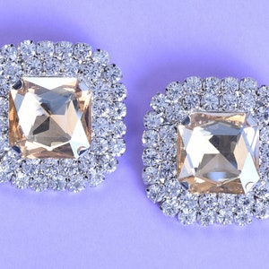 Puede incluir: Dos adornos de strass de forma cuadrada con un centro de cristal transparente y un marco de metal plateado.