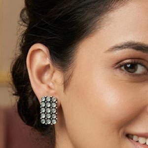 May include: A close-up of a person wearing rectangular stud earrings. The earrings are silver-toned and feature a grid pattern of small, round, light-colored stones. The person is smiling, and a silver necklace is visible.