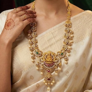 May include: A woman wearing a gold necklace and matching earrings. The necklace features a central pendant with a figure, surrounded by gemstones and pearls. The earrings have a similar design. The woman is wearing a cream-colored sari.