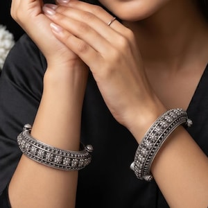 May include: Two silver bangles with detailed floral patterns. The bangles are displayed on the wrists. The jewellery is set against a black backdrop. The bangles appear to be metal and have a vintage or antique aesthetic.