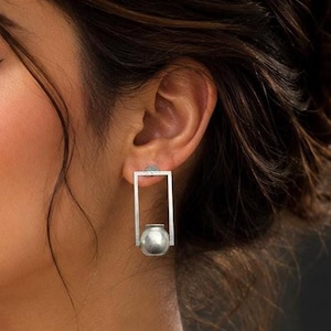 Puede incluir: Un pendiente de tono plateado que presenta un marco rectangular con una cuenta esférica suspendida. El pendiente tiene un diseño geométrico limpio con un acabado metálico, mostrando una estructura de barra rectangular de plata pulida.