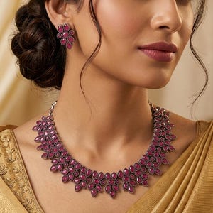 May include: A woman wearing a matching jewelry set. The set includes a necklace, earrings, a ring, and a bracelet. The jewelry features a floral design with dark red gemstones set in a silver-toned metal.