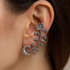 May include: Silver-toned ear cuff featuring a floral design with a crescent moon shape. The cuff wraps around the ear, with a flower at the top and a vine-like structure with small circular accents. The jewelry is worn on the ear.