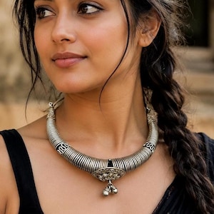 Può includere: Una collana girocollo in metallo color argento caratterizzata da un design a coste e avvolto, con un medaglione decorativo centrale e due piccoli ciondoli pendenti. La collana poggia sul collo, mostrando una finitura metallica e dettagli.