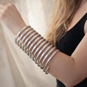 May include: Silver-toned arm cuff featuring a series of curved, parallel bars. The cuff wraps around the forearm, creating a bold, modern look. The jewelry is made of a shiny, metallic material.