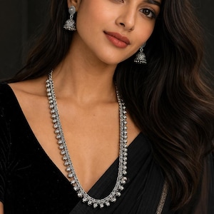 May include: A silver necklace with a long, vertical design, featuring small, round embellishments. The necklace is complemented by matching silver earrings. The model is wearing a black velvet top and a black garment.