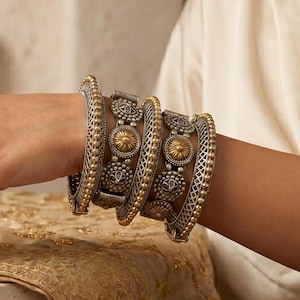 May include: Ornate silver and gold bangles with intricate floral and geometric designs. The bangles are stacked on a wrist, showcasing the detailed craftsmanship and contrasting metal tones. The jewelry is set against a neutral background, highlighting the accessories.