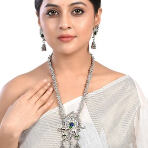 May include: A silver necklace with a peacock pendant featuring a blue and green eye, green accents, and a silver flute. The pendant is attached to a silver chain with small silver beads. The necklace is worn by a person with dark hair and a white top.