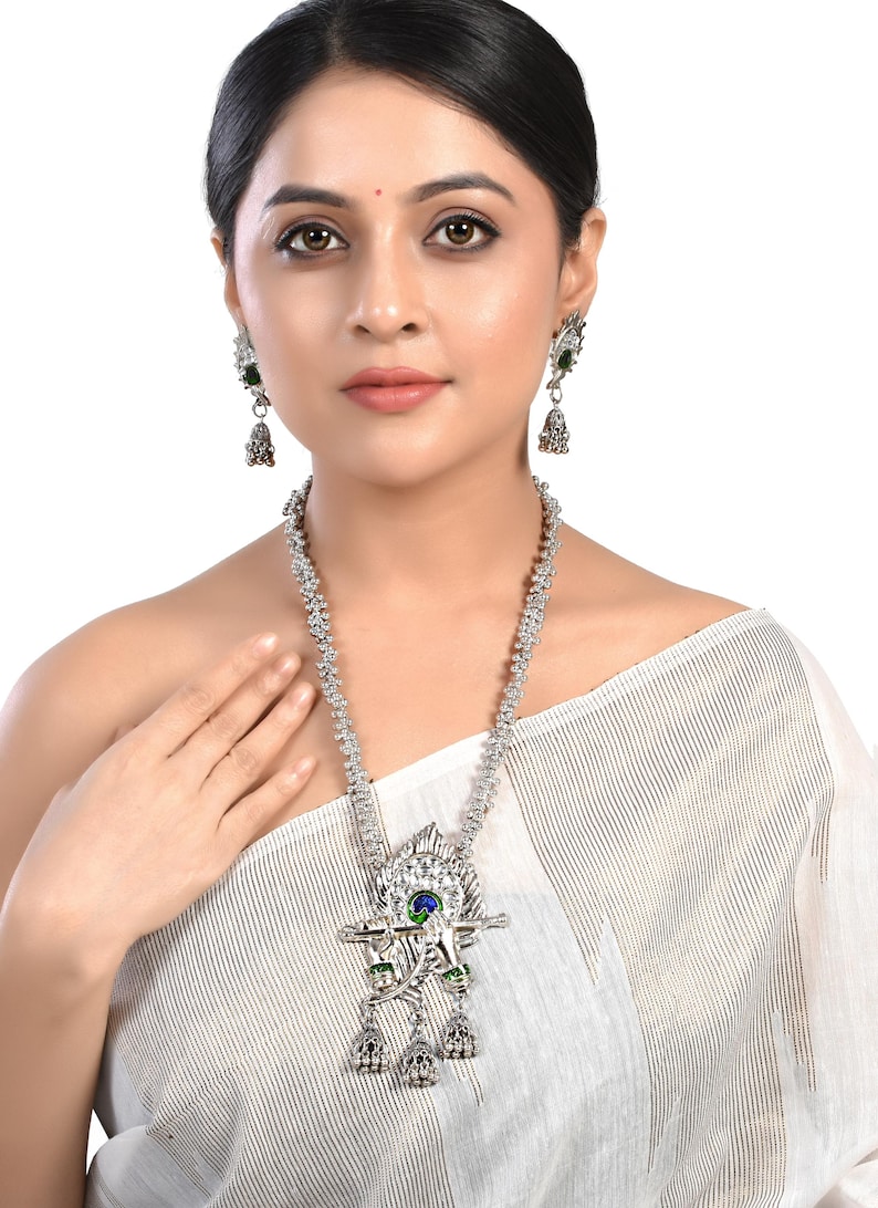 May include: A silver necklace with a peacock pendant featuring a blue and green eye, green accents, and a silver flute. The pendant is attached to a silver chain with small silver beads. The necklace is worn by a person with dark hair and a white top.