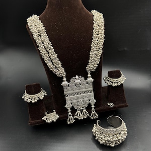 Op de afbeelding: Een zilverkleurige sieradenset met een lange ketting met een grote, sierlijke hanger, twee kleine ringen met hangende bedels en een armband met een brede band en hangende bedels.