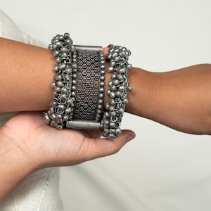 Könnte beinhalten: Ein Set aus vier silbernen Armbändern mit komplizierten Designs. Die Armbänder sind auf dem Handgelenk einer Person übereinander gelegt und weisen eine Kombination aus strukturiertem Metall und baumelnden Perlen auf.