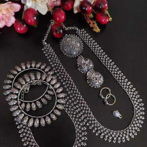 Op de afbeelding: Een zilverkleurige sieradenset met een lange ketting, een paar oorbellen, een neusring en een paar armbanden. De ketting heeft een delicate ketting met een bloemmotief en een grote hanger. De oorbellen zijn ook bloemvormig en de armbanden zijn simpel en rond.