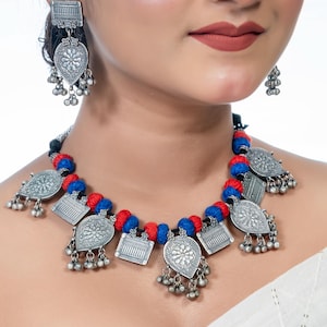 Könnte beinhalten: Eine silberne Halskette mit blauen und roten Fäden und silbernen Anhänger-Anhängern. Die Anhänger-Anhänger sind tropfenförmig und haben komplizierte Designs. Die Halskette wird von einer Person mit einem weißen Oberteil getragen.