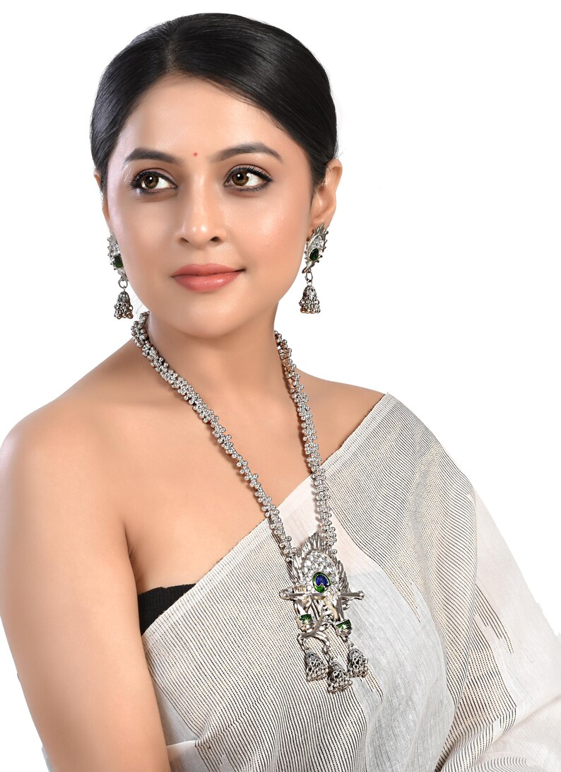 May include: A silver necklace with a large pendant featuring a peacock design with green and blue stones. The necklace is paired with matching earrings with green and blue stones.