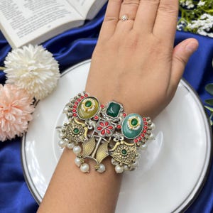 Puede incluir: Una pulsera plateada ornamentada con múltiples detalles de piedras preciosas. La pulsera presenta piedras verdes y amarillas ovaladas, cuadradas y redondas, detalles rojos y perlas de imitación. El diseño incluye filigranas doradas y está sobre un plato blanco.