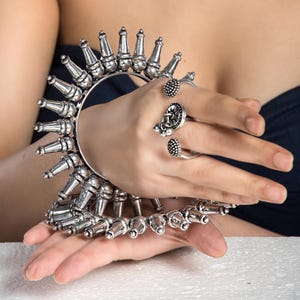 Może przedstawiać: Srebrna bransoletka z ozdobnym wzorem kolców wokół krawędzi. Bransoletka jest noszona na nadgarstku. Srebrny pierścionek z ozdobnym wzorem jest noszony na palcu.