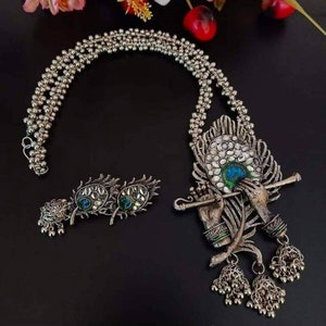 May include: A silver necklace with a peacock design pendant featuring a figure playing a flute. The pendant has blue, green, and white enamel accents. The necklace also includes a pair of matching earrings with peacock feather designs.