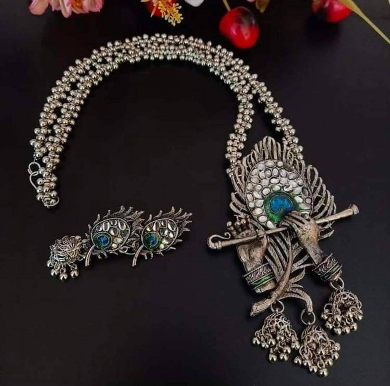 May include: A silver necklace with a peacock design pendant featuring a figure playing a flute. The pendant has blue, green, and white enamel accents. The necklace also includes a pair of matching earrings with peacock feather designs.