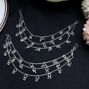 May include: Two silver anklets with multiple strands of delicate chain. Each chain is adorned with small, dangling charms. The anklets are displayed on a black surface, with a glimpse of a white and gold plate and flowers in the background.