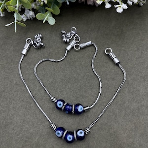 May include: Silver-toned anklet and bracelet set. The anklet and bracelet feature a chain design with dark blue beads, each with a white and blue eye design. The set includes decorative charms.