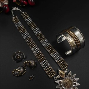 May include: A collection of silver and gold-toned jewelry on a black surface. The set includes a necklace with a sunburst pendant, a cuff bracelet, earrings, and a nose ring. The necklace and bracelet have intricate designs.