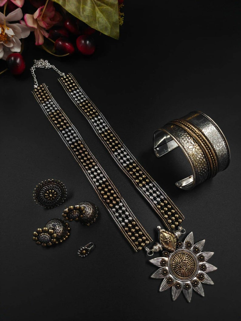 May include: A collection of silver and gold-toned jewelry on a black surface. The set includes a necklace with a sunburst pendant, a cuff bracelet, earrings, and a nose ring. The necklace and bracelet have intricate designs.