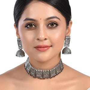 Könnte beinhalten: Ein oxidiertes Silber-Choker-Halsband und Ohrring-Set mit filigranen Blumendesigns. Die Halskette hat eine quadratische Form und die Ohrringe sind im Jhumka-Stil.
