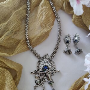 May include: Silver necklace with a peacock feather pendant featuring a depiction of Krishna playing the flute. The pendant has blue and green enamel accents. The necklace is paired with matching earrings.