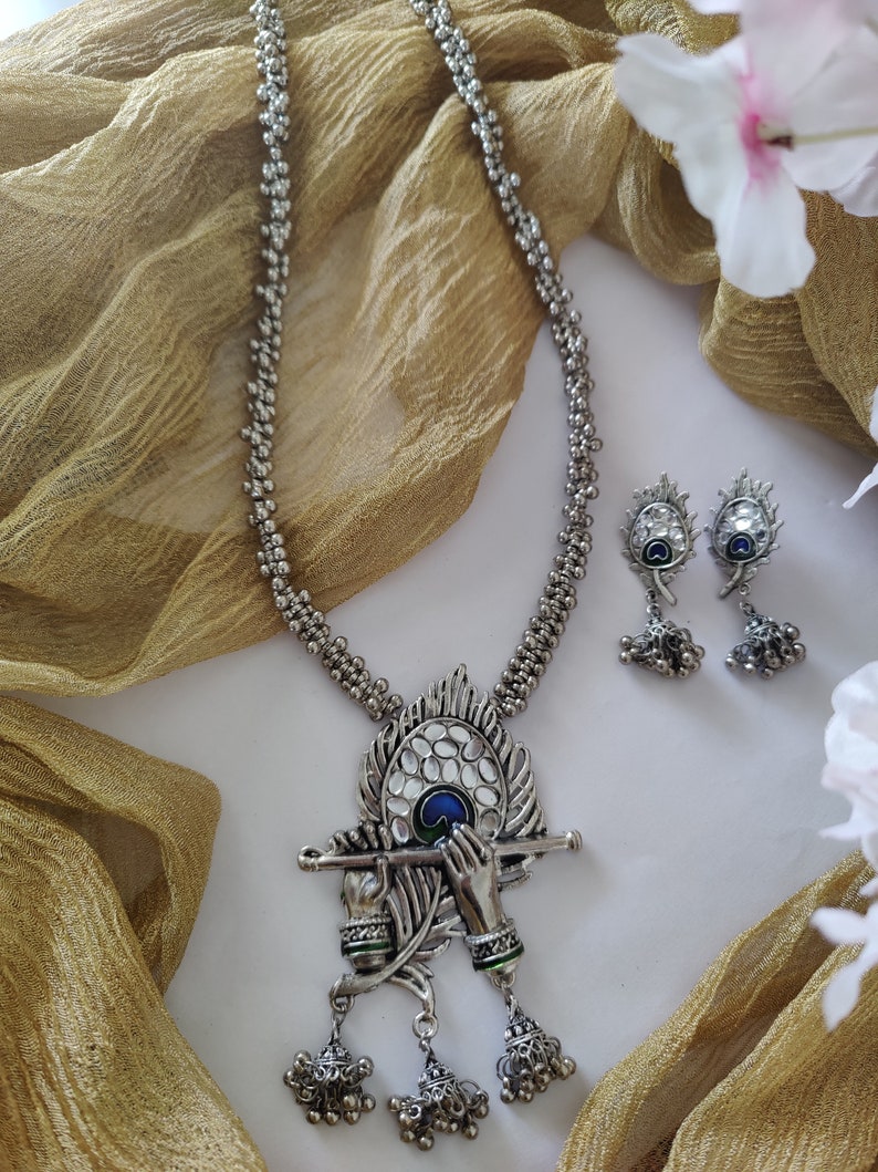 May include: Silver necklace with a peacock feather pendant featuring a depiction of Krishna playing the flute. The pendant has blue and green enamel accents. The necklace is paired with matching earrings.