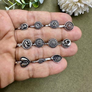 Può includere: Un set di dieci anelli per il naso in argento con diversi design, tra cui un cuore, una foglia, un fiore e un pavone. Gli anelli sono esposti su una mano.