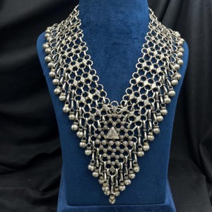 May include: A silver-toned necklace with a geometric design. The necklace features a series of diamond-shaped links and small, dangling bell-shaped charms. The central pendant has a triangular design. The necklace is displayed on a blue mannequin.
