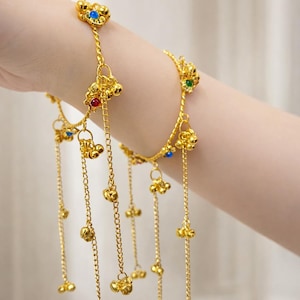 May include: A gold hand chain bracelet with dangling chains and small bell charms. The bracelet features decorative floral elements with blue, red, and green gemstones. The jewelry is worn on a wrist.