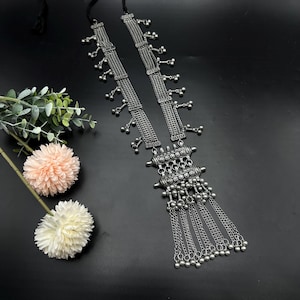 May include: A silver tone long necklace with a multi-layered chain design and a large pendant with a geometric pattern. The pendant has a fringe of smaller chains with silver beads.