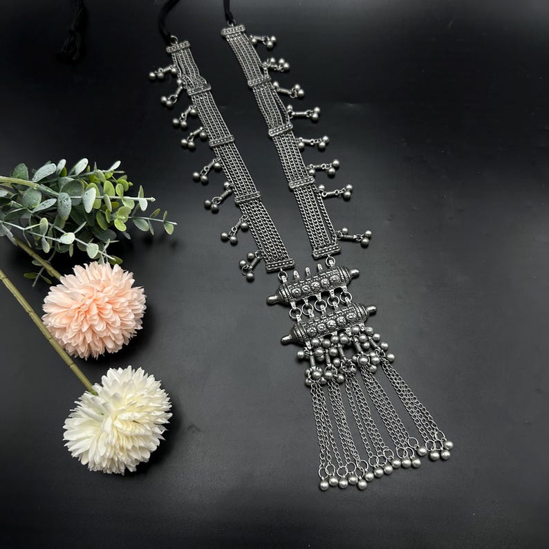 May include: A silver tone long necklace with a multi-layered chain design and a large pendant with a geometric pattern. The pendant has a fringe of smaller chains with silver beads.
