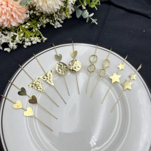 May include: A collection of gold-colored ear climbers with various designs, including hearts, circles, triangles, infinity symbols, and stars. The earrings are arranged on a white plate, showcasing their different shapes and textures.