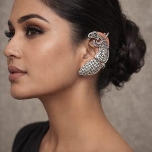 Puede incluir: Un ear cuff plateado con un diseño ornamentado. El ear cuff envuelve la oreja y tiene un patrón detallado con un motivo de pájaro. La joya se lleva en la oreja y es una pieza llamativa.