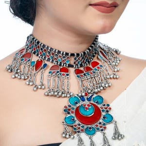 May include: A silver-toned statement necklace with red, blue, and turquoise enamel accents. The necklace features a choker-style design with dangling elements and a large pendant with a floral motif. The jewelry is likely handmade.