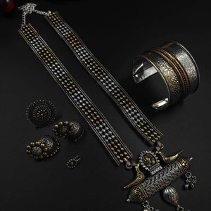 May include: An ornate silver and gold-toned jewelry set on a black surface. The set includes a necklace, cuff bracelet, rings, and earrings. The necklace features a detailed pendant with bird and floral motifs. The cuff bracelet and rings have intricate patterns.