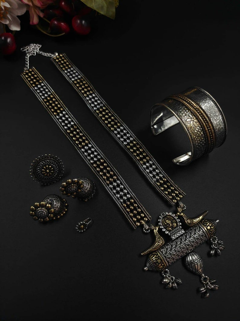 May include: An ornate silver and gold-toned jewelry set on a black surface. The set includes a necklace, cuff bracelet, rings, and earrings. The necklace features a detailed pendant with bird and floral motifs. The cuff bracelet and rings have intricate patterns.