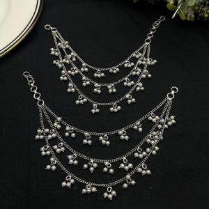 May include: Two silver ear chains, each featuring three tiers of fine chains embellished with small, white, pearl-like beads. The chains connect to a hook and a clasp, designed to be worn with earrings. The background is black.