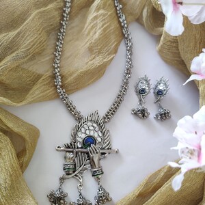 May include: A silver necklace with a peacock feather pendant featuring a pair of hands holding a flute. The pendant has a blue and green enamel design. The necklace is paired with matching earrings.