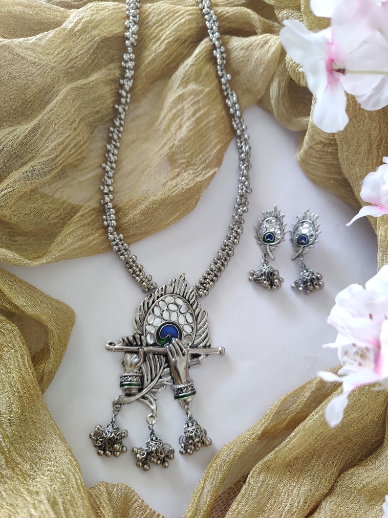 May include: A silver necklace with a peacock feather pendant featuring a pair of hands holding a flute. The pendant has a blue and green enamel design. The necklace is paired with matching earrings.