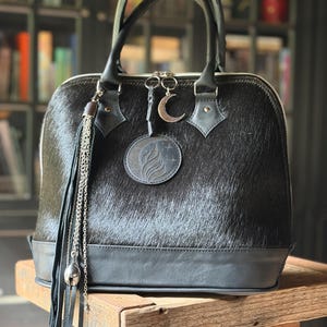 Könnte beinhalten: Eine schwarze Handtasche aus Fell mit glattem Lederboden und Henkeln. Die Tasche verfügt über einen runden Aufnäher mit Flammen- und Sternenmotiv, einen silbernen Mondsichel-Anhänger sowie eine lange schwarze Lederquaste mit Silberkette und Glöckchen.