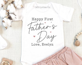 May include: White baby onesie with the text "Happy First Father's Day Love, Evelyn". The onesie is paired with a pink ruffled diaper cover.